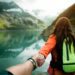 Person holding hands with someone off frame, wearing a green packback, a lake and mountains in the background