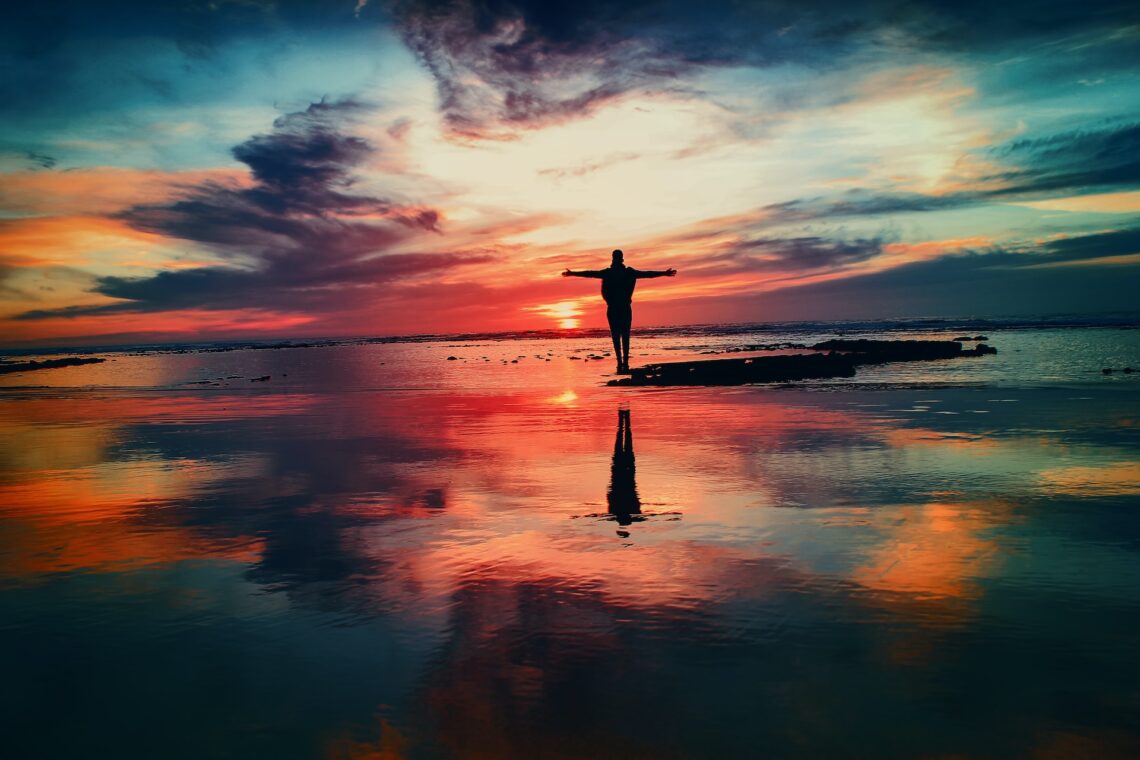 Colourful image of person standing in water, looking at sunset with their arms spread